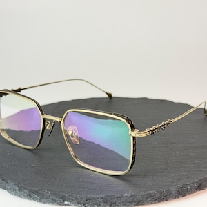 Könnte beinhalten: Goldgerahmte Brille mit quadratischen Gläsern. Die Fassungen haben ein dekoratives Design an den Bügeln und einen schwarzen Akzent um die Gläser. Die Gläser reflektieren ein Regenbogenspektrum. Die Brille liegt auf einer dunklen, grauen, runden Oberfläche.