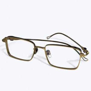 May include: Gold-toned eyeglasses with a rectangular frame and clear lenses. The glasses have a double bridge and a sleek, modern design. The temples are thin and curve around the ears. The overall aesthetic is sophisticated and stylish.