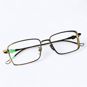 May include: A pair of eyeglasses with a gold-colored metal frame and rectangular lenses. The temples and bridge are thin, and the lenses are clear. The glasses are set against a white background.