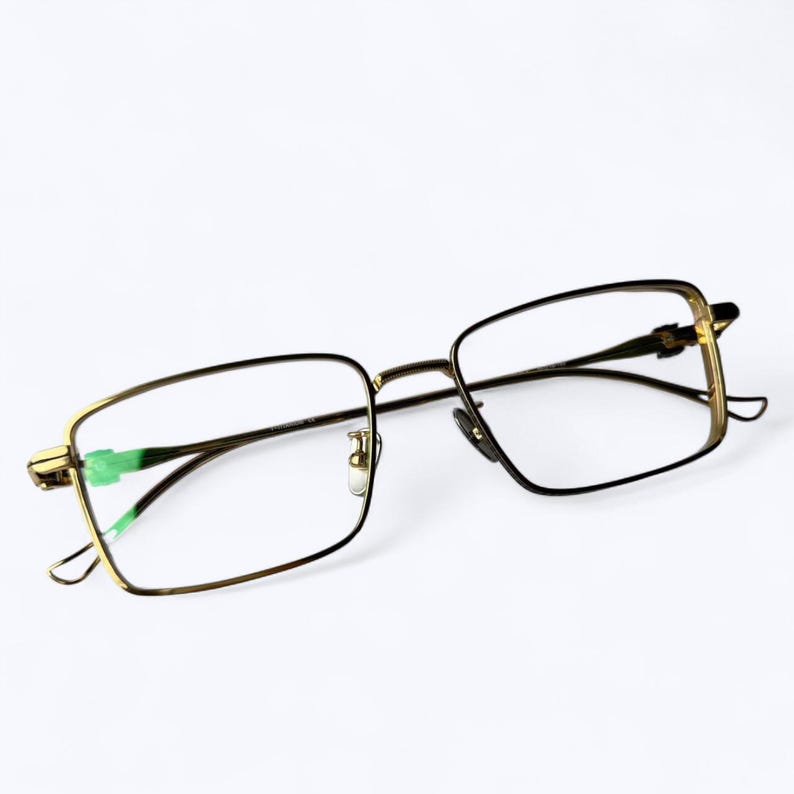 May include: A pair of eyeglasses with a gold-colored metal frame and rectangular lenses. The temples and bridge are thin, and the lenses are clear. The glasses are set against a white background.