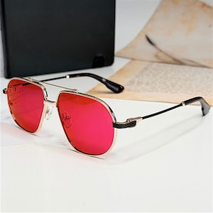 Puede incluir: Gafas de sol de aviador con montura dorada y lentes rojas vibrantes. Las gafas tienen un doble puente y varillas negras con un patrón de carey cerca de las bisagras. El diseño es elegante y moderno.