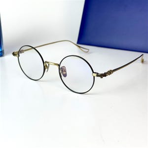 Op de afbeelding: Ronde bril met zwarte montuur en goudkleurige metalen pootjes. De bril heeft een klassiek ontwerp met een brug en neuspads. De lenzen zijn helder en de algehele stijl is vintage.