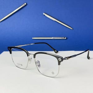 Può includere: Occhiali argento e neri con design a mezza montatura. Le montature hanno una sezione inferiore argentata e una superiore nera. Le aste sono sottili e scure, con dettagli decorativi. Tre penne d'argento sullo sfondo.
