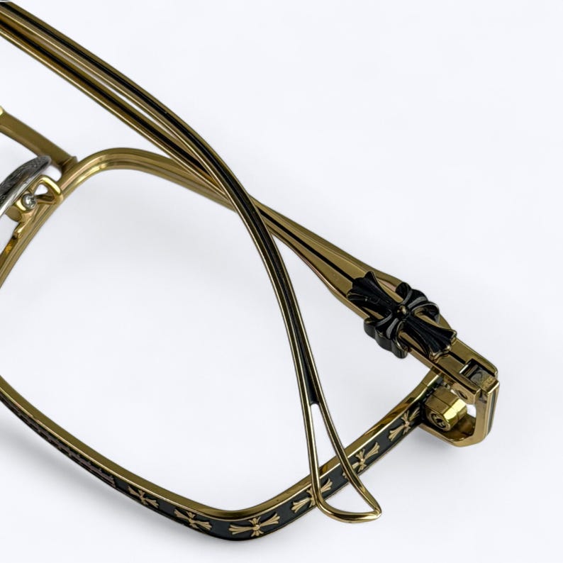 May include: Gold-toned eyeglasses with a black cross design on the temples and frame. The glasses have a rectangular shape and a sleek, modern design. The arms of the glasses are thin and elegant, with a black accent.