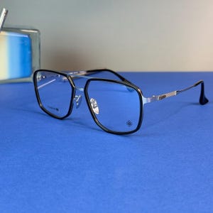 May include: Square-framed eyeglasses with black rims and silver temples. The lenses are clear, and the bridge is silver. The glasses have a double bridge design and a small decorative emblem on the lower right corner of the frame. The background is a solid blue.