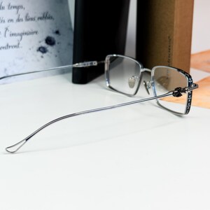 May include: Silver-framed eyeglasses with a rectangular lens shape. The temples have a decorative pattern and the lenses have a gradient tint. The glasses are positioned on a white surface.