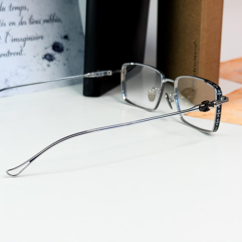 May include: Silver-framed eyeglasses with a rectangular lens shape. The temples have a decorative pattern and the lenses have a gradient tint. The glasses are positioned on a white surface.