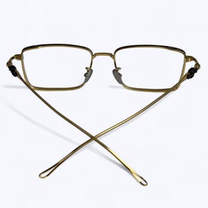 May include: Gold-toned rectangular eyeglasses with clear lenses. The frames have a delicate design, with a thin bridge and temples that cross at the back. The temples have decorative accents near the hinges.