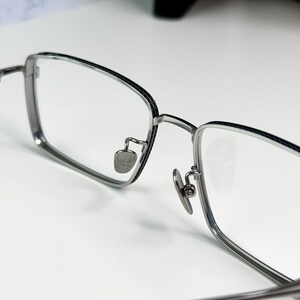 May include: Close-up of eyeglasses with a silver metal frame and clear lenses. The rectangular frames have a thin black accent along the top edge. The nose pads are small and silver. The glasses are on a white surface.