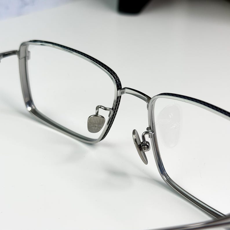 May include: Close-up of eyeglasses with a silver metal frame and clear lenses. The rectangular frames have a thin black accent along the top edge. The nose pads are small and silver. The glasses are on a white surface.