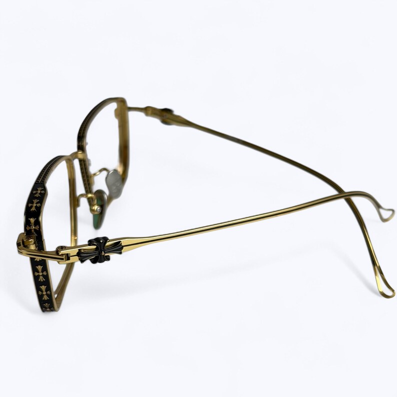 May include: Gold-toned metal eyeglasses with a black accent on the frame and temples. The temples feature a repeating cross pattern. The glasses have a classic, elegant design.