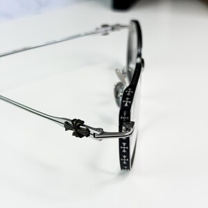 May include: Silver-framed eyeglasses with a black accent on the rims and temples. The temples feature a decorative cross design. The glasses have a classic, elegant style.