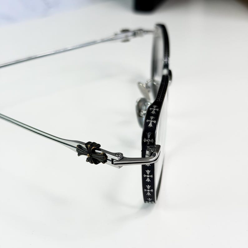 May include: Silver-framed eyeglasses with a black accent on the rims and temples. The temples feature a decorative cross design. The glasses have a classic, elegant style.