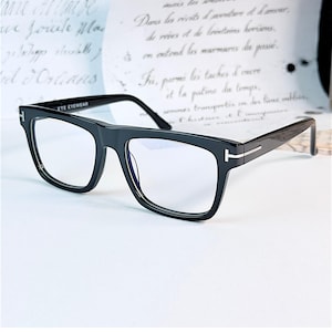 Può includere: Occhiali rettangolari neri con lenti trasparenti e montatura spessa. Le aste presentano un inserto metallico argentato a forma di T sulle cerniere. L'interno dell'asta riporta la scritta ETE EYEWEAR in bianco sulla superficie in plastica nera.