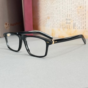 May include: Black rectangular eyeglasses with clear lenses. The frames have gold accents at the temples and small gold rivets. The arms are black with a white and black striped pattern near the hinges. The glasses are set against a neutral background.