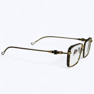 May include: Gold-toned eyeglasses with a rectangular frame and clear lenses. The frame features black detailing with a cross pattern on the temples and hinges. The arms curve gently for a comfortable fit.