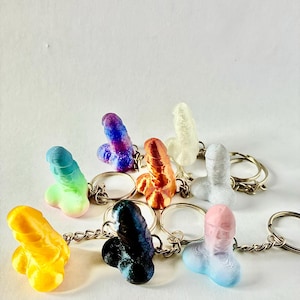 Puede incluir: Un conjunto de ocho llaveros coloridos con forma de parte del cuerpo humano. Los llaveros están hechos de un material liso, similar al plástico, y están disponibles en una variedad de colores, incluyendo azul, verde, naranja, rosa, blanco y morado.
