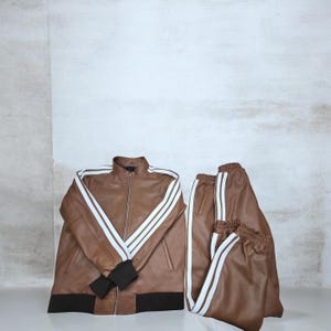 May include: A brown faux leather tracksuit consisting of a zip-up jacket and matching trousers. The jacket features a stand-up collar, black ribbed cuffs and hem, and three white stripes running down each sleeve. The trousers have matching white stripes.