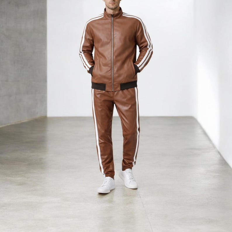 May include: A brown leather tracksuit consisting of a zip-up jacket and matching trousers. The jacket features a high collar, dark brown ribbed cuffs, and two white stripes running down each sleeve. The trousers have matching white side stripes.