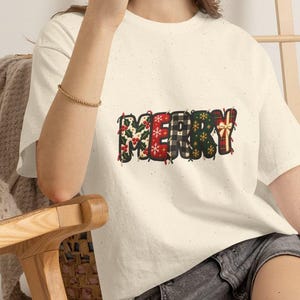 May include: A cream-coloured short-sleeve t-shirt featuring the word MERRY printed across the chest. Each letter has a distinct holiday-themed pattern, including holly leaves, red snowflakes, black and white buffalo plaid, and a gift box design.