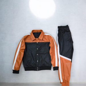 May include: A two-piece athletic set featuring a black and orange jacket and matching pants. The jacket has a black body with an orange yoke and white stripes on the sleeves. The pants are black with orange panels and white stripes.