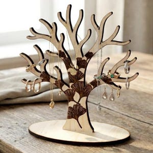 Op de afbeelding: Een houten sieradenboom in de vorm van een boom met takken, met kettingen en ringen eraan. De boom heeft een lichte houtkleur met een donkerdere, patroonstam. Hij staat op een houten tafel bij een raam en een houten kom.