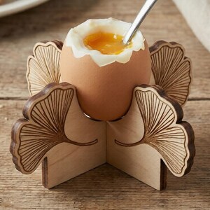 May include: A soft-boiled egg in a wooden egg cup with a ginkgo leaf design. The egg has a runny yolk and is being spooned. The egg cup is made of light-coloured wood with darker etched details. The background is a wooden surface.