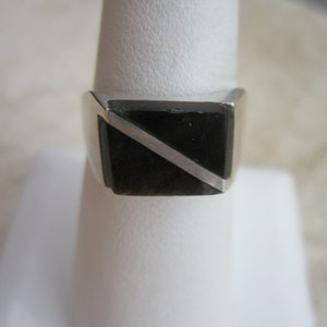 May include: A silver ring with a rectangular black stone, bisected by a diagonal silver stripe. The ring is displayed on a white, cylindrical stand, showcasing its design and craftsmanship. The ring's band is a simple, polished silver.