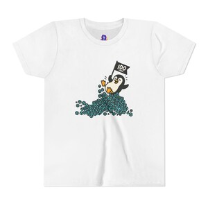 May include: White t-shirt featuring a cartoon penguin holding a black flag with the number 100. The penguin is emerging from a pile of blue cubes. The shirt has a crew neck and short sleeves.