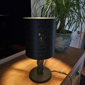 Op de afbeelding: Een zwarte tafellamp met een cilindrische kap bespikkeld met kleine witte stippen. De lamp heeft een donkergrijze voet en steel en staat op een houten oppervlak. Een groene plant in een grijze pot staat op de achtergrond.