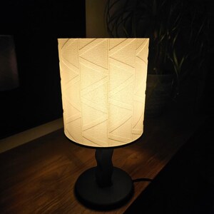 Op de afbeelding: Een kleine, decoratieve tafellamp met een crèmekleurige lampenkap met een geometrisch patroon. De lamp heeft een zwarte voet en steel en staat op een houten oppervlak. De lamp is verlicht en geeft een warme gloed. De lampenkap is ongeveer 15 cm hoog.