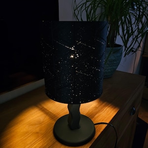 Op de afbeelding: Een zwarte tafellamp met een cilindrische lampenkap met witte stippen en lijnen. De lamp heeft een donkere voet en een gebogen steel. De lamp staat op een houten oppervlak, met een plant op de achtergrond.