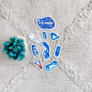 May include: A collection of blue and white stickers featuring medical themes, including a stethoscope, a vial, a diaper, a pregnant figure, an IV bag, and a blood tube, alongside a larger sticker with the text L+D NURSE and medical icons.