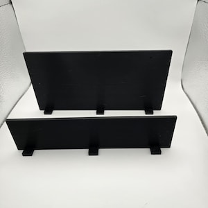 May include: Two black wooden display shelves. The shelves are rectangular and have small, rectangular supports attached to the bottom. The shelves are arranged one above the other, with the top shelf slightly larger than the bottom shelf.