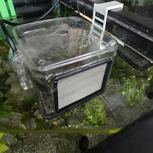 May include: A clear plastic aquarium breeder box with a black-framed mesh side panel, featuring a white plastic mounting bracket attached to the top edge of a glass fish tank, with a black power cord and a green plastic component visible nearby.