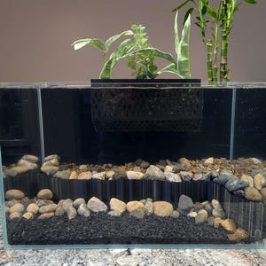 May include: A rectangular glass aquarium filled with water, featuring a black plastic planter basket suspended at the top containing green plants. The bottom is layered with black gravel and various smooth, multi-colored river stones.