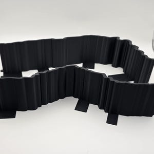 May include: Two black, wavy-edged landscape edging sections. Each section has a series of vertical ridges and small tabs for ground anchoring. The pieces are made of a solid, matte black material.