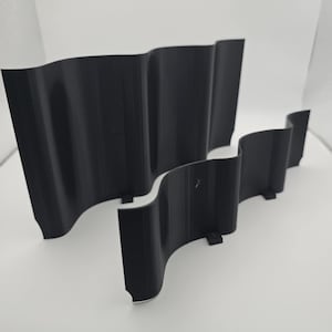 May include: Two black, wavy, 3D-printed objects. The objects have a curved design, resembling a stylized flag or wave. They are placed on a white surface, with a simple, modern aesthetic. The objects appear to be made of plastic.