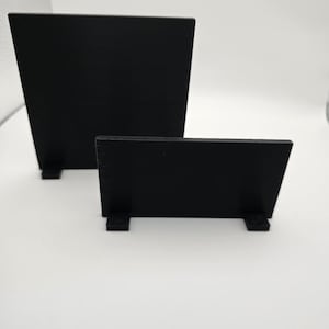 May include: Two black wooden display stands. One is taller and wider than the other. Both have a rectangular shape with a slanted top and a small base for stability. The stands are set against a white background.