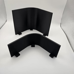 May include: Two black, curved corner protectors. Each protector has a right-angle shape with a flat base and a curved top. The protectors are made of a solid, matte black material and are placed on a white surface.