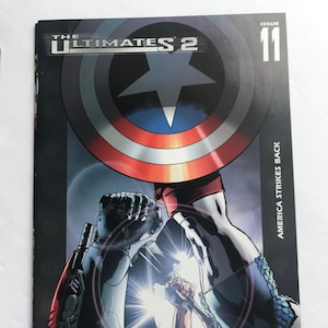 Könnte beinhalten: Comic-Cover für The Ultimates 2, Ausgabe 11. Das Cover zeigt eine Nahaufnahme von Captain America und einer Roboterfigur, wobei Captain Americas Faust in die Luft erhoben ist. Das Cover ist rot, weiß und blau, mit dem Titel "The Ultimates 2" und "Ausgabe 11" in weißem Text. Der Text "America Strikes Back" ist ebenfalls auf dem Cover zu sehen.