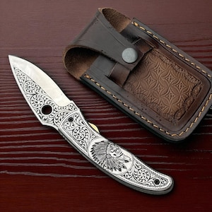 May include: A silver folding knife with intricate scrollwork and an image of a Native American chief. The knife has a partially open blade and rests next to a brown leather sheath with a button closure.