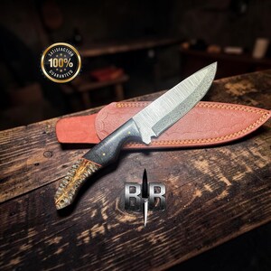 Puede incluir: Un cuchillo de hoja fija con una hoja de metal estampada y un mango que presenta una sección oscura y una sección de grano de madera texturizada de color claro. El cuchillo descansa sobre una funda de cuero beige con costuras visibles. Sello 100%.