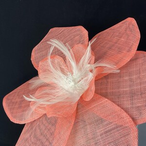 K&ouml;nnte beinhalten: Eine gro&szlig;e, korallenfarbene Blume aus Netzstoff mit einem wei&szlig;en Federschmuck. Die Blume ist ein Dekorationsst&uuml;ck f&uuml;r einen Hut oder ein anderes Accessoire.
