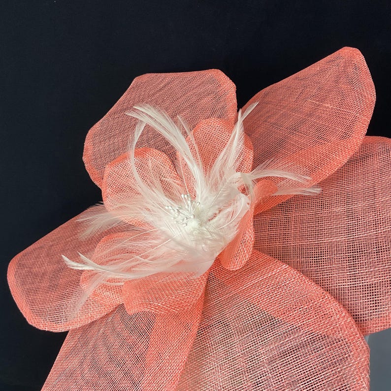 K&ouml;nnte beinhalten: Eine gro&szlig;e, korallenfarbene Blume aus Netzstoff mit einem wei&szlig;en Federschmuck. Die Blume ist ein Dekorationsst&uuml;ck f&uuml;r einen Hut oder ein anderes Accessoire.