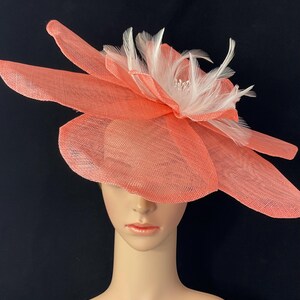 K&ouml;nnte beinhalten: Ein korallenfarbener Fascinator mit einem gro&szlig;en Blumendesign und wei&szlig;en Federn. Der Fascinator ist aus einem zarten, gewebten Material gefertigt.