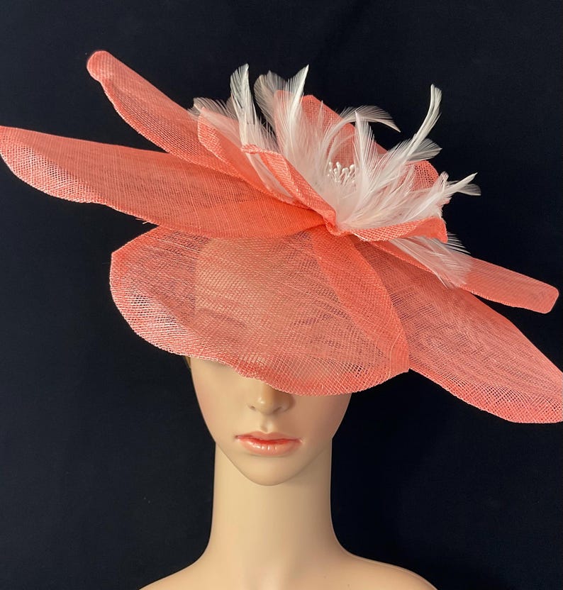 K&ouml;nnte beinhalten: Ein korallenfarbener Fascinator mit einem gro&szlig;en Blumendesign und wei&szlig;en Federn. Der Fascinator ist aus einem zarten, gewebten Material gefertigt.