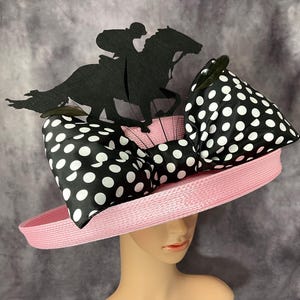 Puede incluir: Un sombrero de paja rosa con un lazo de lunares blanco y negro y una silueta negra de un jinete montando un caballo en la parte superior.