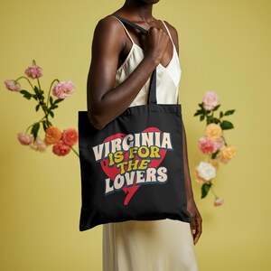 May include: Black tote bag with the text "Virginia is for Lovers" in a retro font, set against a red heart graphic. The bag has black straps and is held by a person. The background is a solid yellow with floral accents.