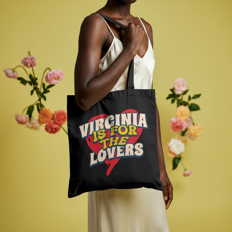 May include: Black tote bag with the text "Virginia is for Lovers" in a retro font, set against a red heart graphic. The bag has black straps and is held by a person. The background is a solid yellow with floral accents.